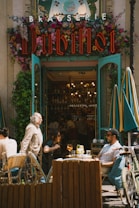 A lively street scene in front of a brasserie entrance adorned with vibrant flowers and neon signage. People are seated at outdoor tables, enjoying drinks while others are walking by. The atmosphere is bustling and warm, emphasized by the open doors revealing a glimpse of the cozy interior with a well-stocked bar.