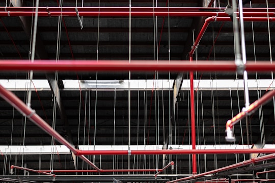 An intricate network of red pipes and metal rods is suspended from the ceiling within a dark industrial or warehouse environment. The pipes are interconnected, supported by thin metal cables that hang vertically from above.