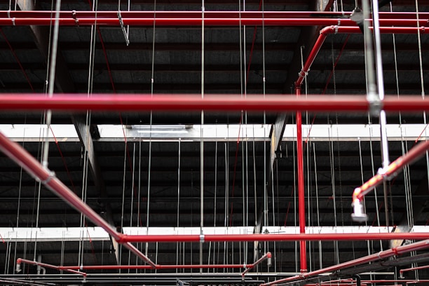 An intricate network of red pipes and metal rods is suspended from the ceiling within a dark industrial or warehouse environment. The pipes are interconnected, supported by thin metal cables that hang vertically from above.