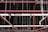 An intricate network of red pipes and metal rods is suspended from the ceiling within a dark industrial or warehouse environment. The pipes are interconnected, supported by thin metal cables that hang vertically from above.