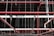 An intricate network of red pipes and metal rods is suspended from the ceiling within a dark industrial or warehouse environment. The pipes are interconnected, supported by thin metal cables that hang vertically from above.