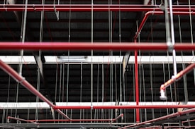 An intricate network of red pipes and metal rods is suspended from the ceiling within a dark industrial or warehouse environment. The pipes are interconnected, supported by thin metal cables that hang vertically from above.