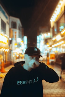 A person is standing in a brightly lit, bustling urban street at night. The street is adorned with numerous neon lights and signs, creating a vibrant atmosphere. The person is wearing a black hoodie with the word 'ESSENTIALS' and a black cap, partially covering their face with their hand.