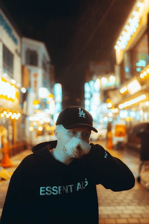 A person is standing in a brightly lit, bustling urban street at night. The street is adorned with numerous neon lights and signs, creating a vibrant atmosphere. The person is wearing a black hoodie with the word 'ESSENTIALS' and a black cap, partially covering their face with their hand.