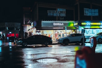 A nighttime street scene featuring a brightly lit clothing store named 'Banilla' and a convenience store called 'CU.' Several parked cars line the street, and the wet pavement reflects the city lights. Overhead signs with Korean writing are visible, adding an urban feel to the setting.