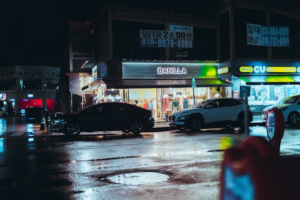 A nighttime street scene featuring a brightly lit clothing store named 'Banilla' and a convenience store called 'CU.' Several parked cars line the street, and the wet pavement reflects the city lights. Overhead signs with Korean writing are visible, adding an urban feel to the setting.