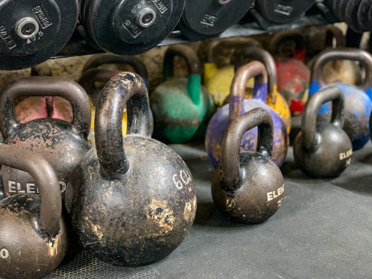 A collection of kettlebells in various sizes and colors is arranged on a gym floor. Some weights have noticeable wear, showcasing their frequent use. The background includes dumbbells on a rack.