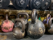 A collection of kettlebells of varying sizes and weights placed on a textured surface. In the background, there is a rack of weight plates. The kettlebells have a worn, rustic appearance with visible markings and a range of colors, including pink, black, and yellow.
