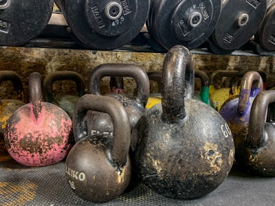 A collection of kettlebells of varying sizes and weights placed on a textured surface. In the background, there is a rack of weight plates. The kettlebells have a worn, rustic appearance with visible markings and a range of colors, including pink, black, and yellow.