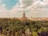 A grand architectural structure with a central tower surrounded by lush greenery. The building has ornate features and seems historic, blending harmoniously with the urban backdrop. The sky above is partly cloudy, casting a soft light over the scene.