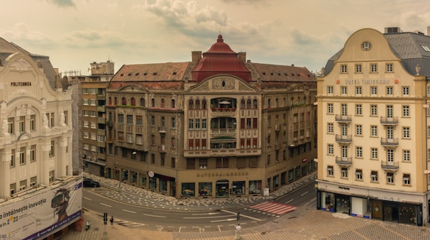 A cityscape featuring historic European architecture, with a beige building displaying 'Politehnica' on the left and 'Hotel Timisoara' on the right. The central building has a red roof and ornate details. The streets are mostly empty except for a few pedestrians.