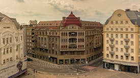A cityscape featuring historic European architecture, with a beige building displaying 'Politehnica' on the left and 'Hotel Timisoara' on the right. The central building has a red roof and ornate details. The streets are mostly empty except for a few pedestrians.