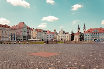 A vibrant and historic town square is depicted, surrounded by colorful, ornate buildings with classic European architecture featuring multiple roof shapes and designs. The foreground consists of a large cobblestone plaza, and the background showcases a partly cloudy sky. Street lamps and sparse people can be seen adding a touch of liveliness to the serene ambiance.
