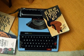 A vintage typewriter with a light blue casing sits on an orange carpet. Several books surround the typewriter, including one prominently titled 'Her Head Was a Spider's Nest' resting on top of it. The books have distinct, artistic covers with bold typography.