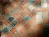 A close-up view of a stone tile floor with a diamond pattern composed of various shades of orange, beige, and grey tiles. The tiles appear worn and textured, with some visible dirt or stains, and they are arranged diagonally.