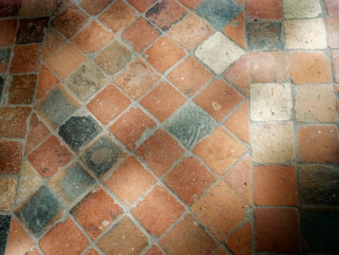 A close-up view of a stone tile floor with a diamond pattern composed of various shades of orange, beige, and grey tiles. The tiles appear worn and textured, with some visible dirt or stains, and they are arranged diagonally.