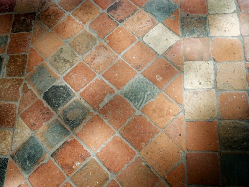 A close-up view of a stone tile floor with a diamond pattern composed of various shades of orange, beige, and grey tiles. The tiles appear worn and textured, with some visible dirt or stains, and they are arranged diagonally.