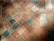 A close-up view of a stone tile floor with a diamond pattern composed of various shades of orange, beige, and grey tiles. The tiles appear worn and textured, with some visible dirt or stains, and they are arranged diagonally.