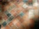 A close-up view of a stone tile floor with a diamond pattern composed of various shades of orange, beige, and grey tiles. The tiles appear worn and textured, with some visible dirt or stains, and they are arranged diagonally.