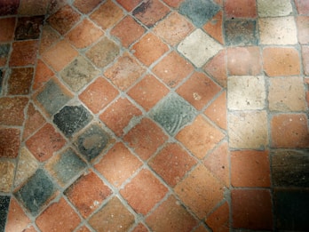 A close-up view of a stone tile floor with a diamond pattern composed of various shades of orange, beige, and grey tiles. The tiles appear worn and textured, with some visible dirt or stains, and they are arranged diagonally.