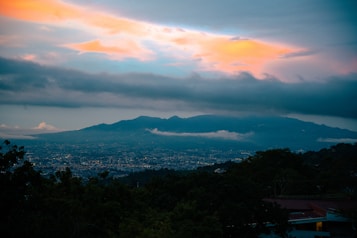 A panoramic view of a city surrounded by lush greenery with a mountain range in the distance. The sky displays a stunning gradient of colors from deep blue to vibrant orange and pink, indicating either sunrise or sunset. Dense clouds partially cover the peaks of the mountains, adding depth to the scene.