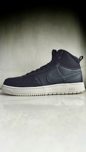 A black high-top sneaker with a prominent white sole is positioned against a plain, textured background. The sneaker has a recognizable brand logo on the side and features eyelets for laces.