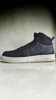 A black high-top sneaker with a prominent white sole is positioned against a plain, textured background. The sneaker has a recognizable brand logo on the side and features eyelets for laces.