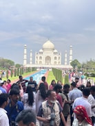 A crowd of people gathers in front of the iconic Taj Mahal, which stands majestically against a cloudy sky. The foreground is bustling with visitors, some taking photos and others engaged in conversation. The reflective pool leads the eye towards the beautiful white marble structure, flanked by well-maintained gardens.