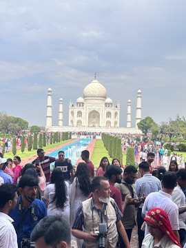 A crowd of people gathers in front of the iconic Taj Mahal, which stands majestically against a cloudy sky. The foreground is bustling with visitors, some taking photos and others engaged in conversation. The reflective pool leads the eye towards the beautiful white marble structure, flanked by well-maintained gardens.