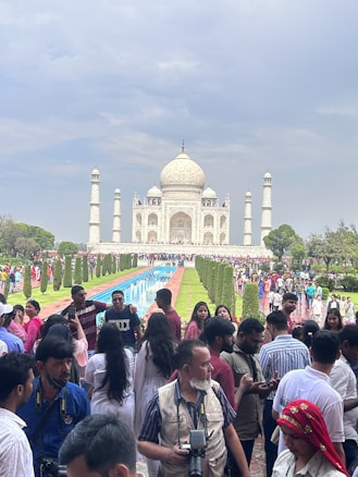 A crowd of people gathers in front of the iconic Taj Mahal, which stands majestically against a cloudy sky. The foreground is bustling with visitors, some taking photos and others engaged in conversation. The reflective pool leads the eye towards the beautiful white marble structure, flanked by well-maintained gardens.