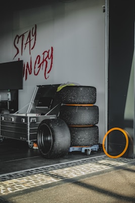 A stack of worn racing tires is placed on a small wheeled cart beside a large metallic storage case. To the side, a single tire is leaning against a wall. Bold red graffiti reading 'STAY HUNGRY' is written on the wall above the tires. The floor is lined with stone tiles, and soft sunlight is casting shadows across the scene.
