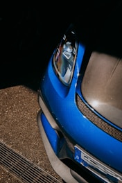 The close-up view of a blue car features a sleek, aerodynamic design. The image focuses on the front section, highlighting the headlight, part of the grille, and bumper. The surrounding area includes a textured asphalt surface, providing a dynamic contrast between the car's glossy finish and the matte ground.