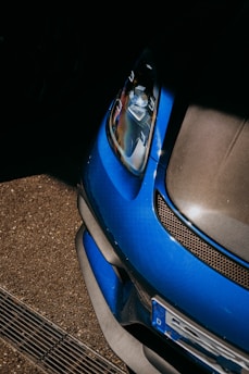The close-up view of a blue car features a sleek, aerodynamic design. The image focuses on the front section, highlighting the headlight, part of the grille, and bumper. The surrounding area includes a textured asphalt surface, providing a dynamic contrast between the car's glossy finish and the matte ground.