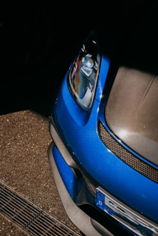 The close-up view of a blue car features a sleek, aerodynamic design. The image focuses on the front section, highlighting the headlight, part of the grille, and bumper. The surrounding area includes a textured asphalt surface, providing a dynamic contrast between the car's glossy finish and the matte ground.