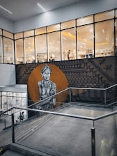 A large mural painted on an exterior wall features a detailed black and white depiction of an elderly indigenous figure with intricate tattoos and traditional attire. The background of the mural is a round, earthy orange circle surrounded by geometric patterns. The mural is located near the entrance of a modern, glass-walled building with visible interiors showcasing people engaged in seated activities.