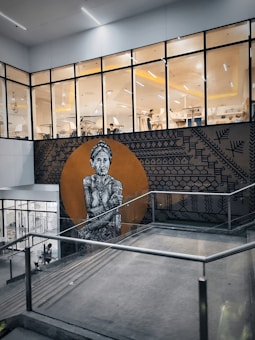 A large mural painted on an exterior wall features a detailed black and white depiction of an elderly indigenous figure with intricate tattoos and traditional attire. The background of the mural is a round, earthy orange circle surrounded by geometric patterns. The mural is located near the entrance of a modern, glass-walled building with visible interiors showcasing people engaged in seated activities.