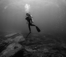 A scuba diver is underwater, surrounded by a vast and serene ocean environment. The diver is equipped with a tank, mask, and fins and is producing bubbles that rise above. The seafloor is visible with rocky formations and a muted texture, suggesting a calm underwater scene.