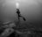 A scuba diver is underwater, surrounded by a vast and serene ocean environment. The diver is equipped with a tank, mask, and fins and is producing bubbles that rise above. The seafloor is visible with rocky formations and a muted texture, suggesting a calm underwater scene.
