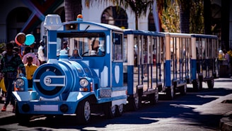 A miniature blue train with a whimsical design travels along a road, surrounded by people carrying colorful balloons. The scene takes place in an outdoor setting with palm trees and buildings in the background. The train appears to be part of a casual, festive environment.
