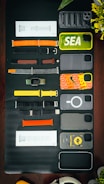 A collection of various smartphone and watch accessories is arranged neatly on a dark surface. The items include colorful phone cases with designs and labels, assorted watch bands in different materials and colors, and some packaged accessories. In addition to the tech accessories, there is a partial view of a green plant with yellow flowers and an over-ear headphone in the corner.