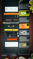 A collection of various smartphone and watch accessories is arranged neatly on a dark surface. The items include colorful phone cases with designs and labels, assorted watch bands in different materials and colors, and some packaged accessories. In addition to the tech accessories, there is a partial view of a green plant with yellow flowers and an over-ear headphone in the corner.