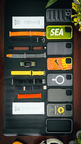 A collection of various smartphone and watch accessories is arranged neatly on a dark surface. The items include colorful phone cases with designs and labels, assorted watch bands in different materials and colors, and some packaged accessories. In addition to the tech accessories, there is a partial view of a green plant with yellow flowers and an over-ear headphone in the corner.