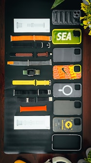 A collection of various smartphone and watch accessories is arranged neatly on a dark surface. The items include colorful phone cases with designs and labels, assorted watch bands in different materials and colors, and some packaged accessories. In addition to the tech accessories, there is a partial view of a green plant with yellow flowers and an over-ear headphone in the corner.