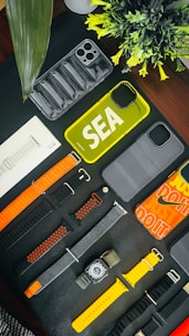 An assortment of phone cases and watch bands are arranged on a dark surface. There are various designs and colors, including yellow and transparent phone cases. The watch bands include orange, black, and yellow hues. A small plant adds a touch of greenery to the scene.