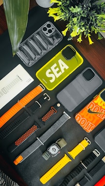 An assortment of phone cases and watch bands are arranged on a dark surface. There are various designs and colors, including yellow and transparent phone cases. The watch bands include orange, black, and yellow hues. A small plant adds a touch of greenery to the scene.
