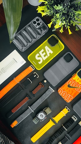 An assortment of phone cases and watch bands are arranged on a dark surface. There are various designs and colors, including yellow and transparent phone cases. The watch bands include orange, black, and yellow hues. A small plant adds a touch of greenery to the scene.