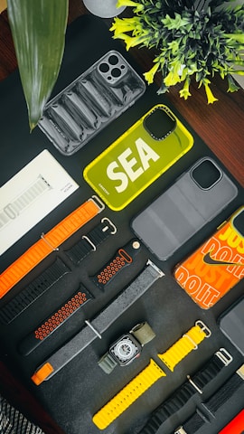 An assortment of phone cases and watch bands are arranged on a dark surface. There are various designs and colors, including yellow and transparent phone cases. The watch bands include orange, black, and yellow hues. A small plant adds a touch of greenery to the scene.