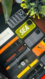 An assortment of phone cases and watch bands are arranged on a dark surface. There are various designs and colors, including yellow and transparent phone cases. The watch bands include orange, black, and yellow hues. A small plant adds a touch of greenery to the scene.
