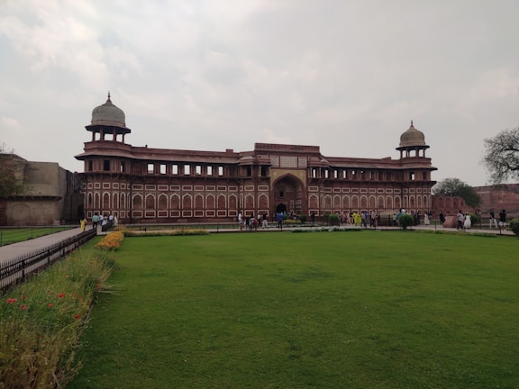 A historic red sandstone building with two domed towers stands prominently at the center of a well-maintained grassy area. Several people are walking around, highlighting its function as a tourist attraction. The sky is mostly cloudy, adding a leisurely ambiance to the scene. Decorative flower beds with vibrant colors line the pathway leading to the entrance.