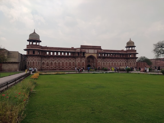 A historic red sandstone building with two domed towers stands prominently at the center of a well-maintained grassy area. Several people are walking around, highlighting its function as a tourist attraction. The sky is mostly cloudy, adding a leisurely ambiance to the scene. Decorative flower beds with vibrant colors line the pathway leading to the entrance.